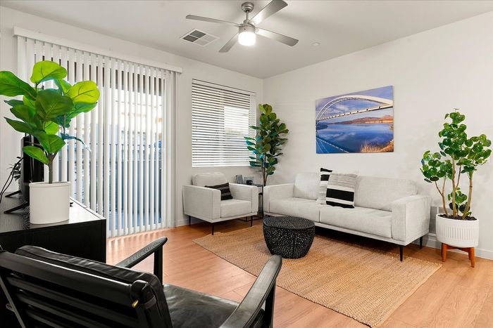 Two-Bedroom Apartments in East Mesa, AZ - Acero Eastmark - Bright Living Room With White Sofa, Accent Chair, Indoor Plants, Wood-Style Flooring, and Glass Doors with Blinds.