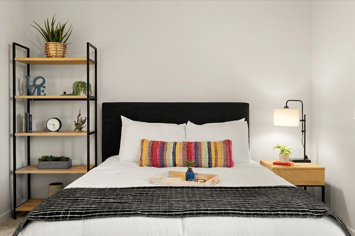 Inviting bedroom with cozy white bedding, colorful accent pillow, and stylish decor including modern shelves and ambient lighting.