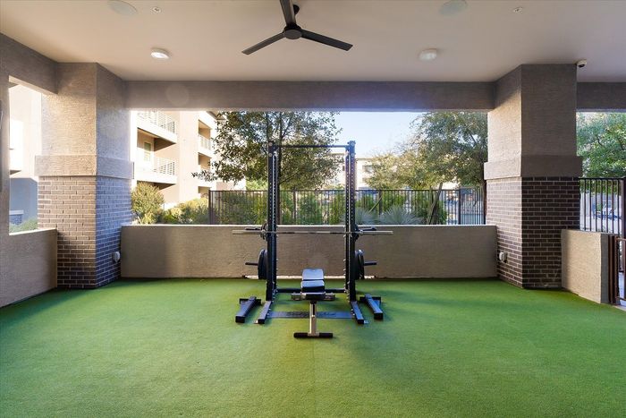 Modern outdoor gym area with state-of-the-art weightlifting equipment on vibrant green artificial turf, surrounded by elegant brickwork and lush greenery.