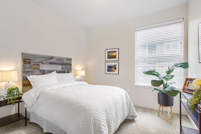 Vancouver, WA, Apartments with In-Unit Washer and Dryer - Acero Jensen Park - A Bedroom with Bedside Tables, Lamps, Wall Art, a Large Window, and Indoor Plants.