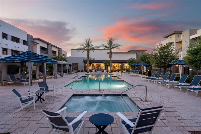 Luxurious apartment complex featuring a stunning pool surrounded by elegant lounge chairs and cabanas, set against a picturesque sunset sky.