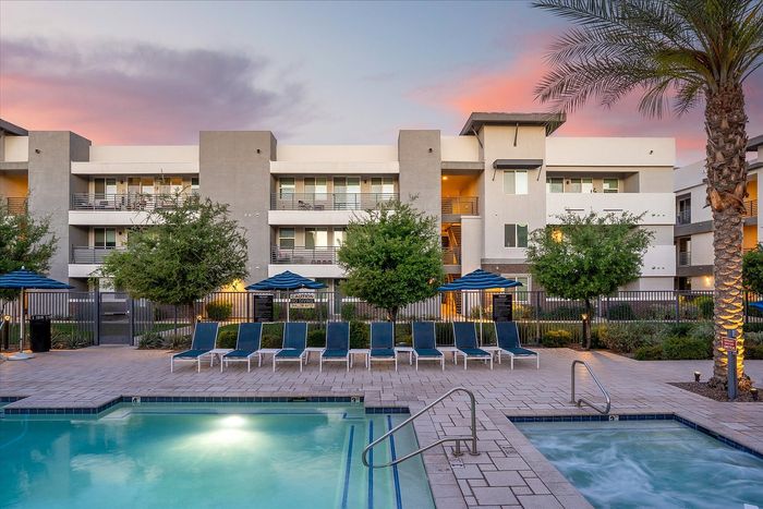 Elegant apartment complex with a sparkling pool, modern design, and palm trees under a vibrant evening sky.