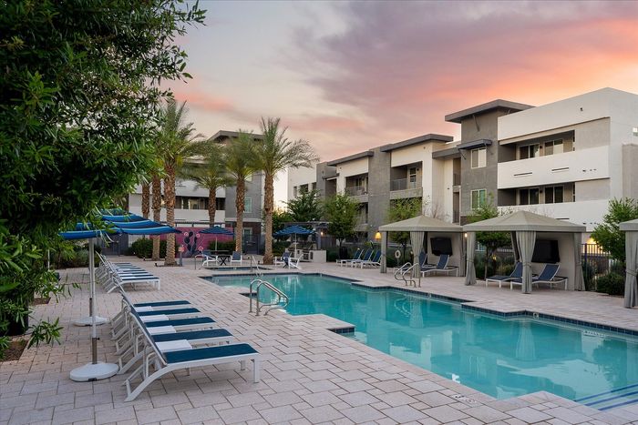 Elegant apartment complex pool area with luxurious cabanas, loungers, and palm trees under a colorful sunset sky.
