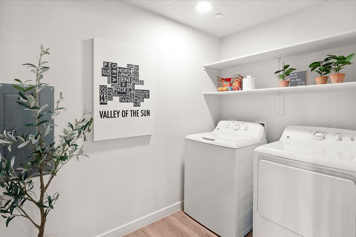 Bright and modern laundry room featuring a washer, dryer, stylish wall art, and shelving with potted plants and storage, ideal for a functional and chic home space.