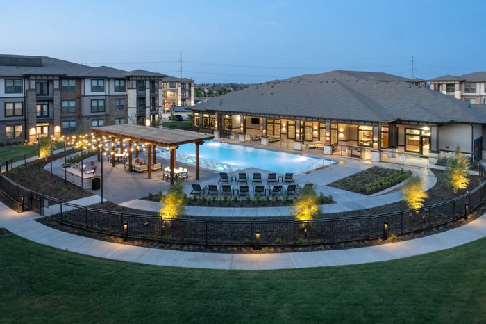 Luxurious apartment complex with a resort-style pool, elegant pergola seating area, and beautifully landscaped grounds at dusk.