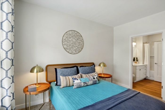The James Apartments | Bedroom with a blue and gray color scheme, featuring a stylish bed with decorative pillows, a round metal wall art piece, and side tables with gold lamps. | Luxury Apartments Haverhill MA