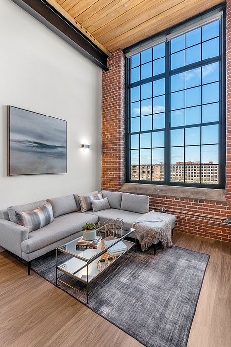 Riverwalk lofts |  A modern living room with exposed brick walls and a large window showing a cityscape. It features a gray sectional sofa, a metallic coffee table with decor, and a gray rug. A framed artwork hangs on the wall, and the ceiling is wooden.| Apartments for rent lawrence ma