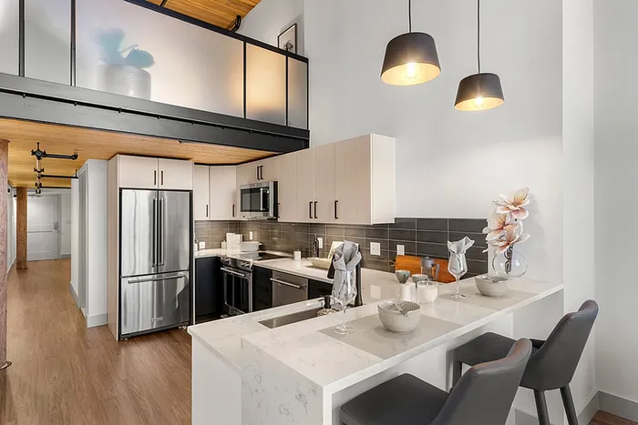 Riverwalk Lofts | Modern loft apartment with a high ceiling and exposed wooden beams. The kitchen features sleek white cabinets and stainless steel appliances, with a breakfast bar and two gray stools. Above, a frosted glass partition hints at an upper level. | Studio Apartments lawrence ma