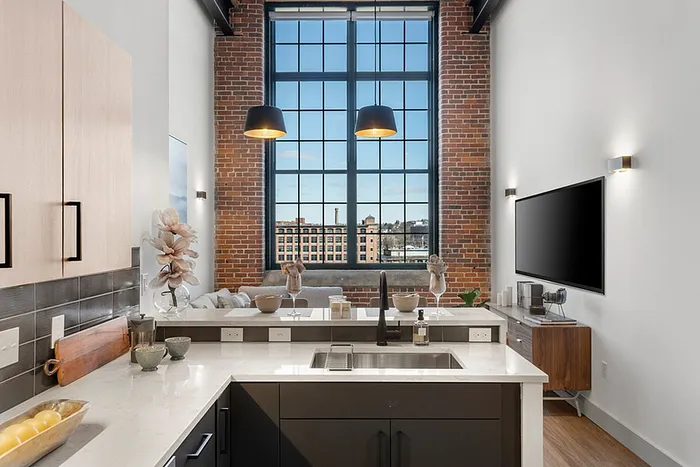 Riverwalk Lofts | A modern loft kitchen with a tall window and exposed brick wall. The kitchen features a white counter, dark cabinets, and pendant lights. A flat-screen TV and decorative items are on the wall, with a cityscape visible through the window. | Loft apartments lawrence ma