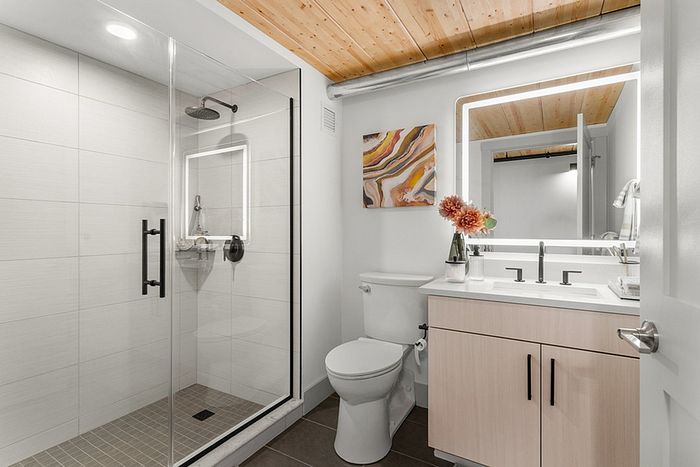 riverwalk lofts | Modern bathroom with a glass-enclosed shower, white tiles, a wooden ceiling, and a light wood vanity. A lit mirror is above the sink. A colorful abstract painting and a vase with flowers are on the vanity. | luxury apartments lawrence ma