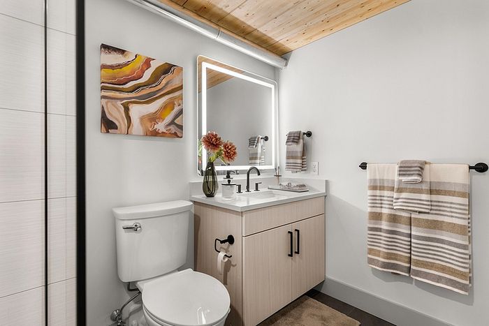 riverwalk lofts | Modern bathroom with a glass-enclosed shower, white tiles, a wooden ceiling, and a light wood vanity. A lit mirror is above the sink. A colorful abstract painting and a vase with flowers are on the vanity. | luxury apartments lawrence ma