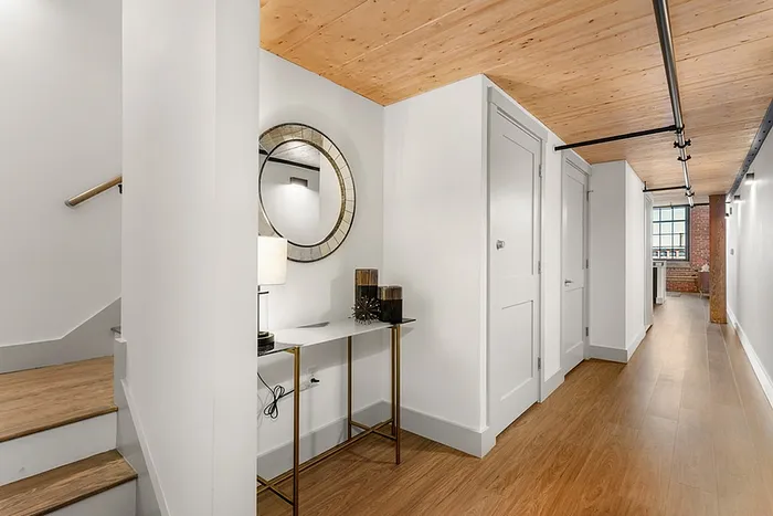 Riverwalk lofts | A modern hallway with light wooden floors and a wooden ceiling. On the left, stairs lead to the next level. A small console table with a lamp and decorative items sits below a round mirror. At the end, a room with a brick wall is visible. | studio apartments for rent ma