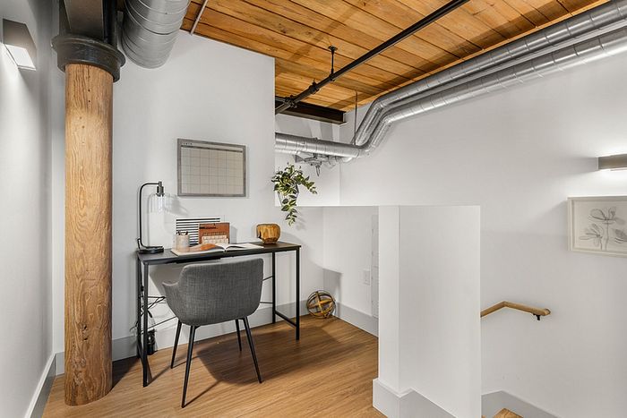 riverwalk lofts | A small modern home office with a desk and chair in a loft area. The space features a wooden column, exposed ductwork, and a wood ceiling. Theres a wall-mounted calendar, a potted plant, and minimal decor, creating a cozy and industrial feel. | Loft apartments lawrence ma