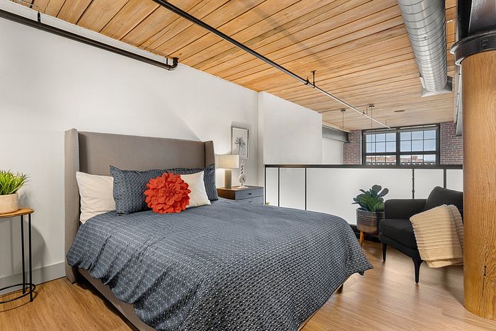riverwalk lofts |  A modern bedroom with a large bed featuring a gray blanket and a red accent pillow. The room has wooden ceilings, a cozy black armchair, a side table with a plant, a large window with brickwork visible outside, and exposed ductwork. | pet friendly apartments lawrence ma