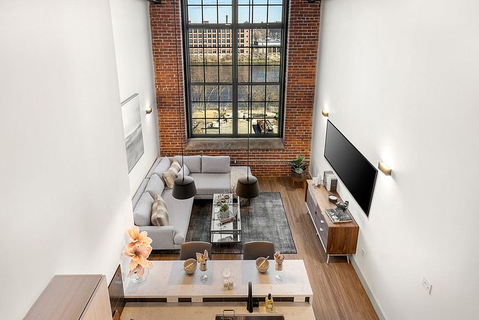 riverwalk lofts | A modern, open-concept loft with large windows showing a cityscape and river. The living area features gray sofas, a TV, and a glass coffee table. The kitchen has a white countertop with bar stools and decorative items. The interior has exposed brick walls.| apartments for rent lawrence ma