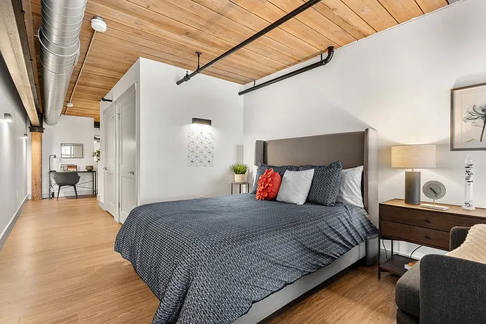 riverwalk lofts | A modern bedroom features a large bed with blue bedding and red accent pillows. A nightstand with a lamp and framed artwork is to the right. The room has wooden floors and a ceiling with exposed beams and ductwork. A desk is in the background. | loft apartments for rent ma