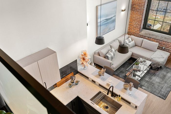 riverwalk lofts | A modern, open-concept loft with large windows showing a cityscape and river. The living area features gray sofas, a TV, and a glass coffee table. The kitchen has a white countertop with bar stools and decorative items. The interior has exposed brick walls.| apartments for rent lawrence ma