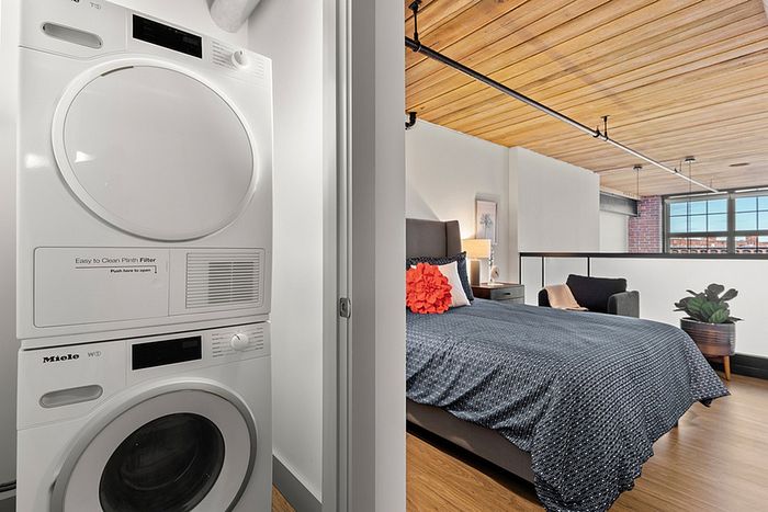 riverwalk lofts | A modern loft features a cozy bedroom with a gray bedspread and a red pillow, adjacent to a laundry area with a stacked washer and dryer. The space is bright, with a wooden ceiling and large windows letting in natural light. | apartments for rent in lawrence ma