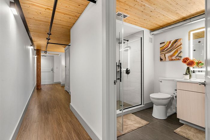 riverwalk lofts | A modern bathroom with a glass-enclosed shower, toilet, and a vanity with a sink. The ceiling is wooden, and theres a hallway with wood flooring. A vase with orange flowers sits on the counter, and a colorful abstract painting hangs on the wall. | luxury apartments massachusetts