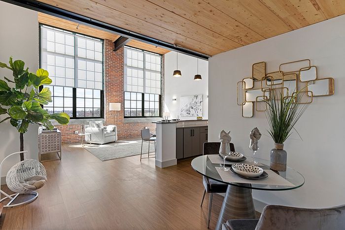 Riverwalk Lofts | A modern, open-concept loft apartment with large windows, exposed brick, and wooden ceilings. The room features a dining area with a glass table, a kitchen with pendant lights, and a cozy seating area. A plant and geometric wall art add decorative touches.| Loft apartments lawrence ma