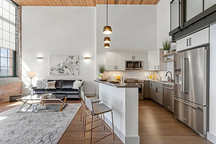 riverwalk lofts | Modern loft apartment with high ceilings, featuring a kitchen with stainless steel appliances and a breakfast bar. The living area has a gray sofa, coffee table, and wall art. Large windows provide abundant natural light. A wooden ceiling adds warmth. | luxury apartments near andover