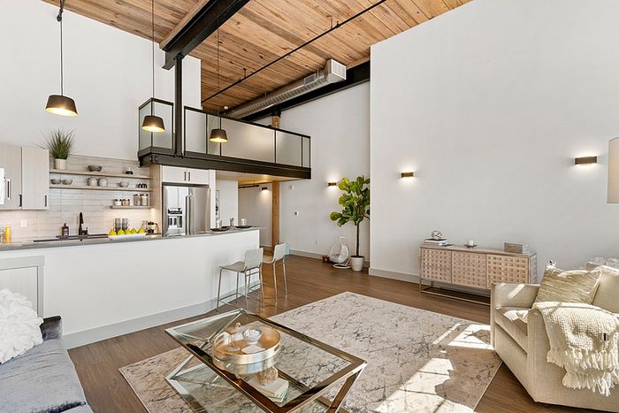 riverwalk lofts | Spacious modern living room with high ceilings and wood accents. Features include a cozy seating area with a glass coffee table, a kitchen with open shelving, a dining area, and a large potted plant. Natural light fills the room from large windows. | one bedroom apartments lawrence ma