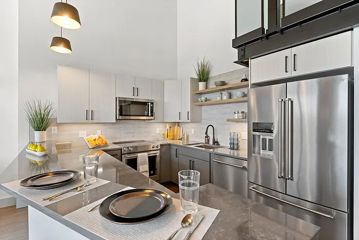 riverwalk lofts | Modern kitchen with stainless steel appliances, grey countertops, and white cabinets. A breakfast bar with black plates and glasses is set for two. Pendant lights hang above, and plants and decor add a touch of greenery. | luxury apartments lawrence ma