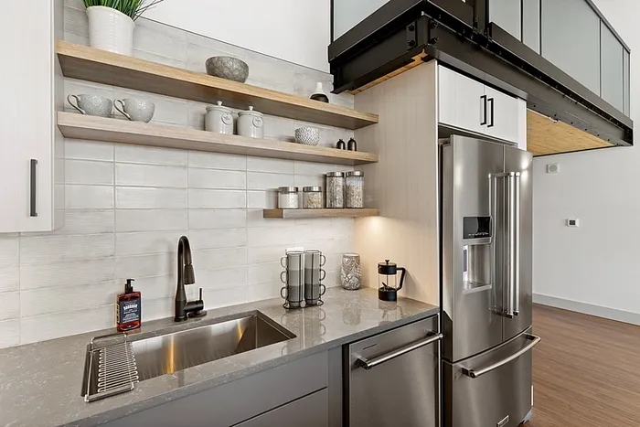 riverwalk lofts | Modern kitchen with light gray cabinetry, a stainless steel refrigerator, and a built-in oven. Open shelves displaying jars and mugs are mounted above the countertop. A black faucet is installed over a large sink, and wooden accents are visible. | open concept apartments lawrence ma