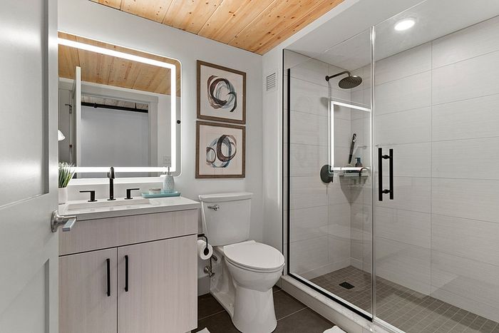 riverwalk lofts | A modern bathroom with a wooden ceiling, featuring a sink with a lighted mirror, a toilet, and a glass-enclosed shower. The walls are adorned with two abstract art pieces, and the overall decor is minimalist and elegant. | one bedroom apartments near boston ma