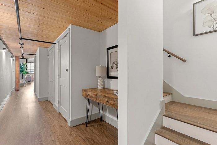 riverwalk lofts | A hallway with light wooden floors and ceiling. On the right, stairs with a wooden handrail lead upwards. A wooden table with a lamp and a framed animal picture decorates the wall. In the distance, a door and a potted plant are visible. | loft apartments lawrence ma