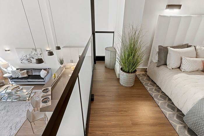 riverwalk lofts | A stylish loft apartment with a view from the upper floor. The bedroom features a bed with neutral bedding, plants, and a rug. Below, a living area has a couch, coffee table, and dining setup. Modern pendant lights hang over both levels. | one bedroom apartments lawrence ma