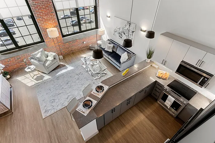 riverwalk lofts | A modern loft apartment features an open-concept living area with large windows, a grey sofa, and a cozy armchair. The kitchen has dark countertops, bar seating, and appliances. The design includes exposed brick walls and light wood flooring. | loft apartments lawrence ma