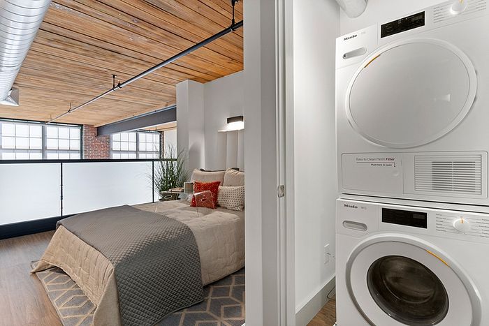 riverwalk lofts | A modern loft features a cozy bedroom with a gray bedspread and a red pillow, adjacent to a laundry area with a stacked washer and dryer. The space is bright, with a wooden ceiling and large windows letting in natural light. | one bedroom apartments for rent near boston
