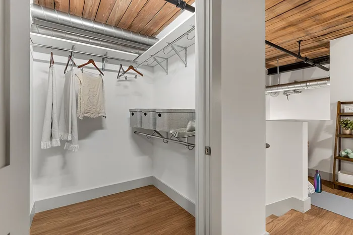 riverwalk lofts | A walk-in closet with a wooden floor and exposed wood ceiling. It contains metal shelving with woven baskets, a few hanging clothes, and a white railing on the right. The space is well-lit and industrial in style. | luxury loft apartments near boston