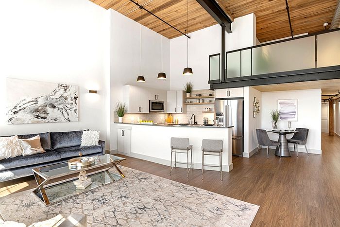 riverwalk lofts | Modern loft apartment with high ceilings, featuring a kitchen with stainless steel appliances and a breakfast bar. The living area has a gray sofa, coffee table, and wall art. Large windows provide abundant natural light. A wooden ceiling adds warmth. | luxury apartments near andover