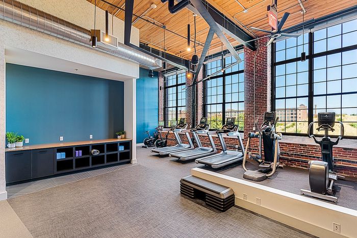 Riverwalk Lofts | A modern gym with large windows, featuring treadmills, stationary bikes, and an exercise step platform. The room has exposed brick walls, a wooden ceiling, and a long cabinet for storage. Bright natural light fills the space. | loft apartments near boston