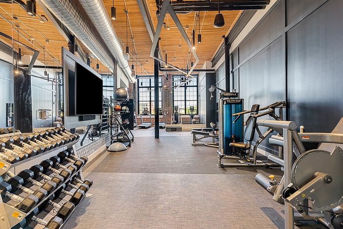 Riverwalk Lofts | A modern gym with various equipment, including dumbbells, weight machines, and cardio machines. The space features large windows, high ceilings with exposed beams, and a mix of natural and artificial lighting. | loft apartments near boston