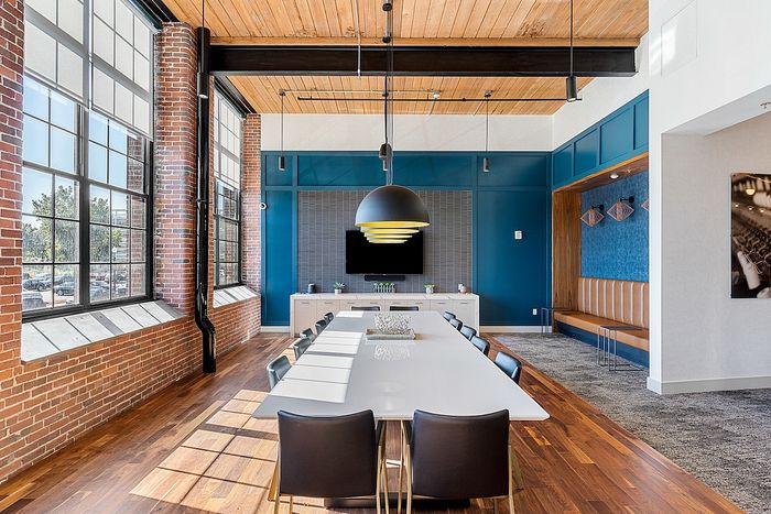 Riverwalk Lofts | A modern conference room with a long white table and black chairs. Large windows with brick frames illuminate the space. Blue accent wall, wooden ceiling, and contemporary lighting fixture enhance the stylish design. | loft apartments near boston