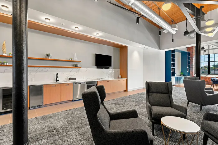 Riverwalk Lofts | A modern lounge area with sleek black armchairs and a small round table on a gray carpet. The background features a wall-mounted TV, a long countertop with a sink, and open shelves displaying decorative items. Natural light fills the space. | luxury apartments near boston