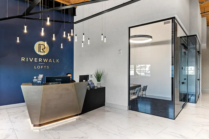 Riverwalk Lofts | A modern office lobby with a large front desk featuring the Riverwalk Lofts logo on a blue wall. Sleek hanging lights illuminate the space. A glass-walled meeting room is visible to the right, with a round table and chairs inside. | luxury apartments near boston