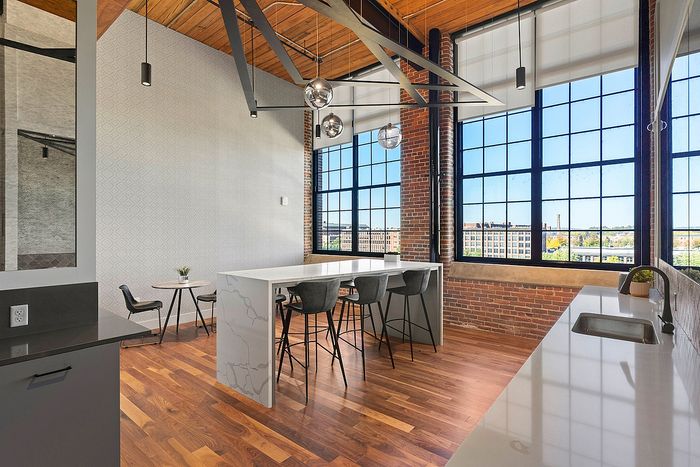 Riverwalk Lofts | A modern kitchen and dining area with high ceilings, featuring a large island with black stools, wooden floors, and industrial-style brick walls. Large windows let in natural light, and a small round table with two chairs is in the corner. | luxury apartments for rent lawrence ma