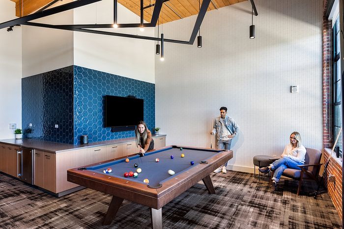 Riverwalk Lofts | A modern game room with three people. One person is playing pool, aiming at the balls on a sleek wooden table. Two others are seated nearby, watching. The room features a wall-mounted TV and stylish decor with blue geometric patterns. | apartments for rent lawrence ma