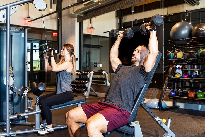 Riverwalk Lofts | Two people are exercising in a gym. A man sits on an inclined bench lifting dumbbells overhead, while a woman uses a cable machine. The gym is equipped with weights and exercise balls in the background. | apartments for rent lawrence ma