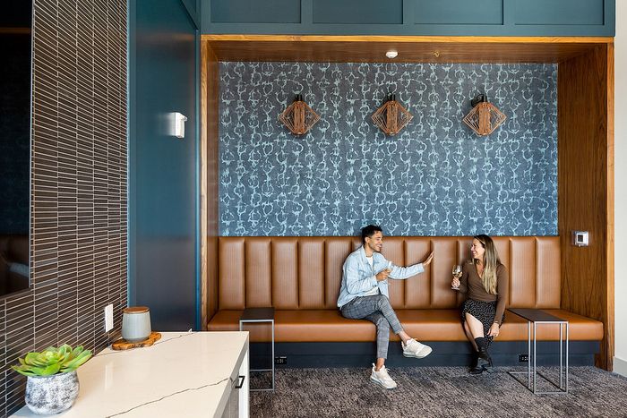 Riverwalk Lofts | A man and woman sit on a brown bench in a modern lounge, clinking glasses and smiling. The room has blue patterned wallpaper, geometric light fixtures, and a countertop with a plant. | apartments for rent lawrence ma