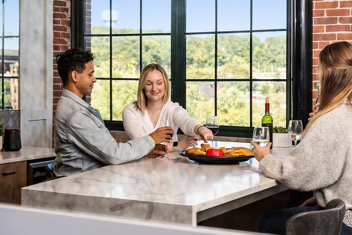 Riverwalk Lofts | Three people sit around a kitchen island with wine glasses, sharing a plate of snacks. Large windows reveal a scenic view of greenery outside. The setting is cozy, with brick walls and a relaxed atmosphere. | apartments for rent lawrence ma