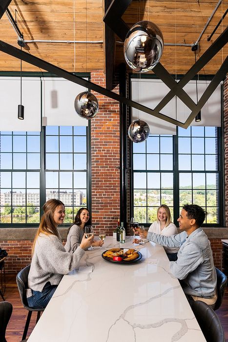 Riverwalk Lofts | Four people sit around a modern dining table with snacks and wine glasses. The room features exposed brick walls, large windows, and contemporary hanging lights. They are smiling and engaged in conversation against a backdrop of a scenic view. | apartments for rent lawrence ma