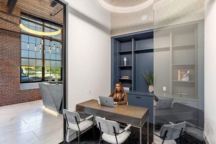 Riverwalk Lofts | A woman sits at a desk in a modern office space with a laptop. The room has a glass wall, blue shelves with decorative items, and a large window showing an industrial-themed lobby with a brick wall and stylish lighting. | luxury apartments for rent lawrence ma