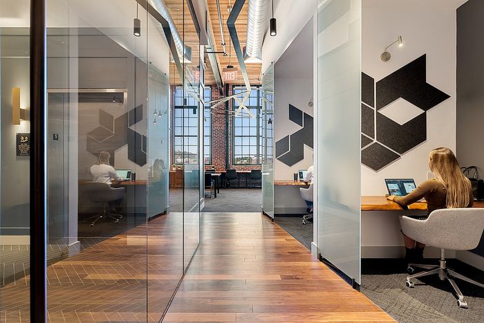 Riverwalk Lofts | A modern office space with glass-walled private cubicles on either side. Two people work on laptops inside separate cubicles. The corridor leads to a communal area with large windows, letting in natural light. Geometric wall art adds a decorative touch. | loft apartments near boston