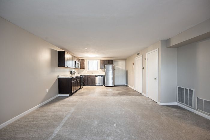 Modern open-plan apartment featuring a sleek kitchen with stainless steel appliances, rich wood cabinetry, and a spacious living area with plush carpeting.