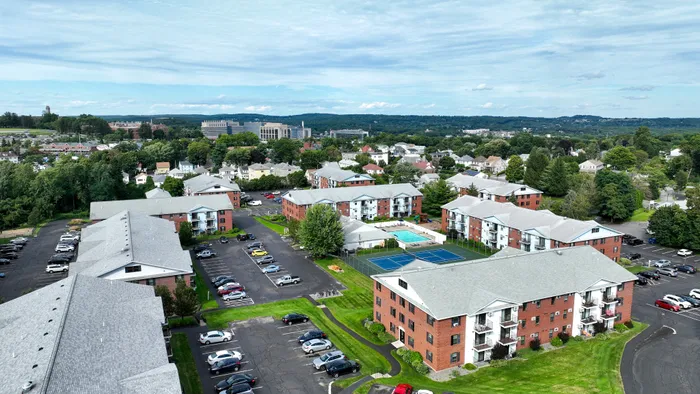 Aerial view of Princeton Place Apartments in Worcester, MA, featuring red brick buildings, a swimming pool, and a tennis court, all surrounded by green lawns, trees, and nearby houses under a partly cloudy sky.